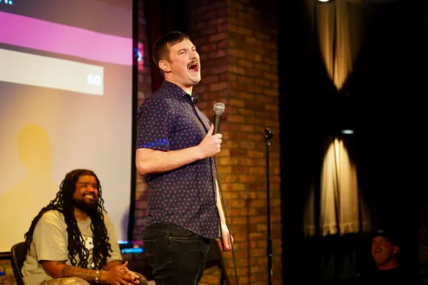 A comedian in a navy patterned shirt performing while another comedian watches from a chair